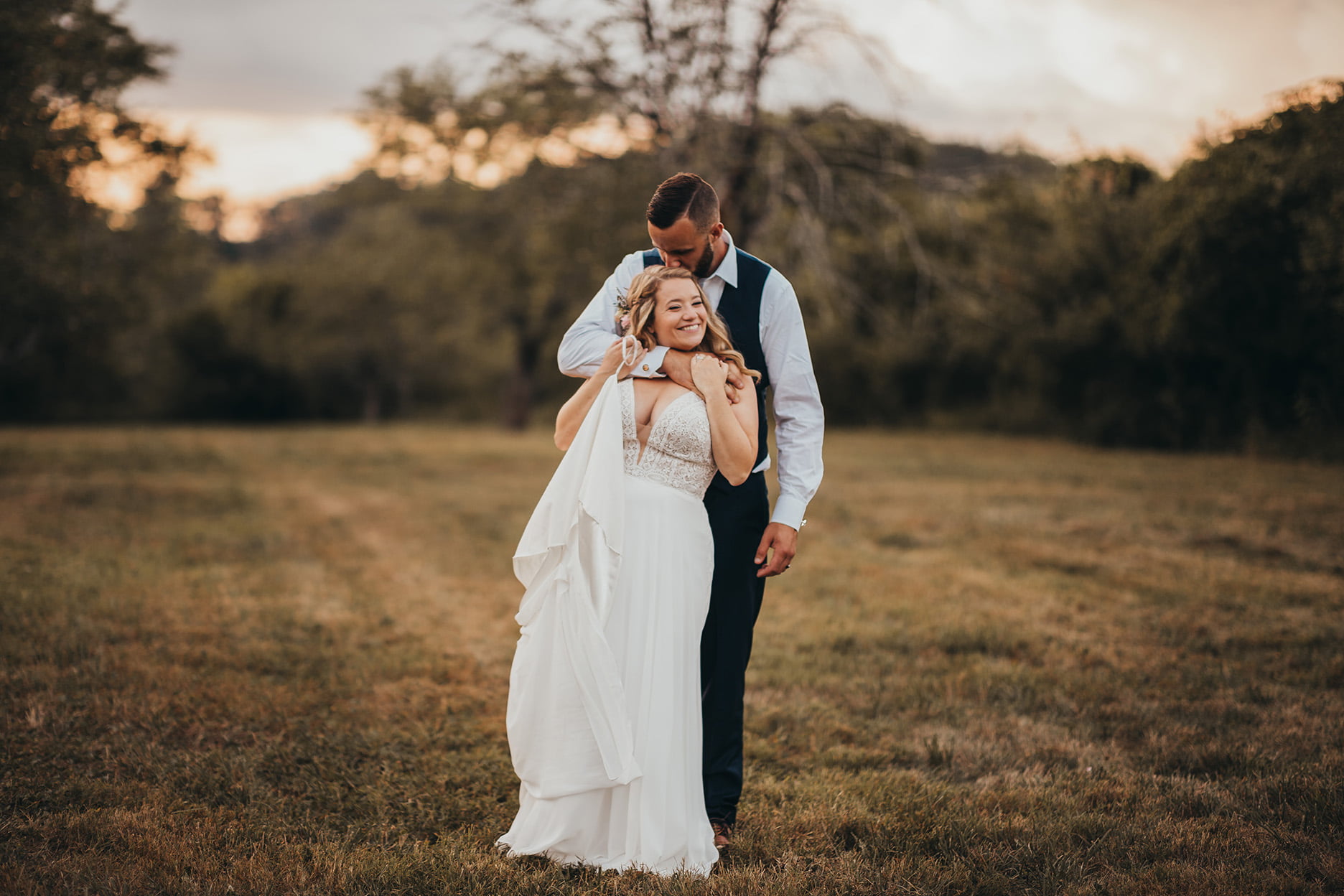A country bride and groom embrace in a field at sunset.