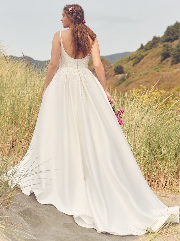 A woman in the Iona wedding dress gracefully walks barefoot on the sandy beach.
